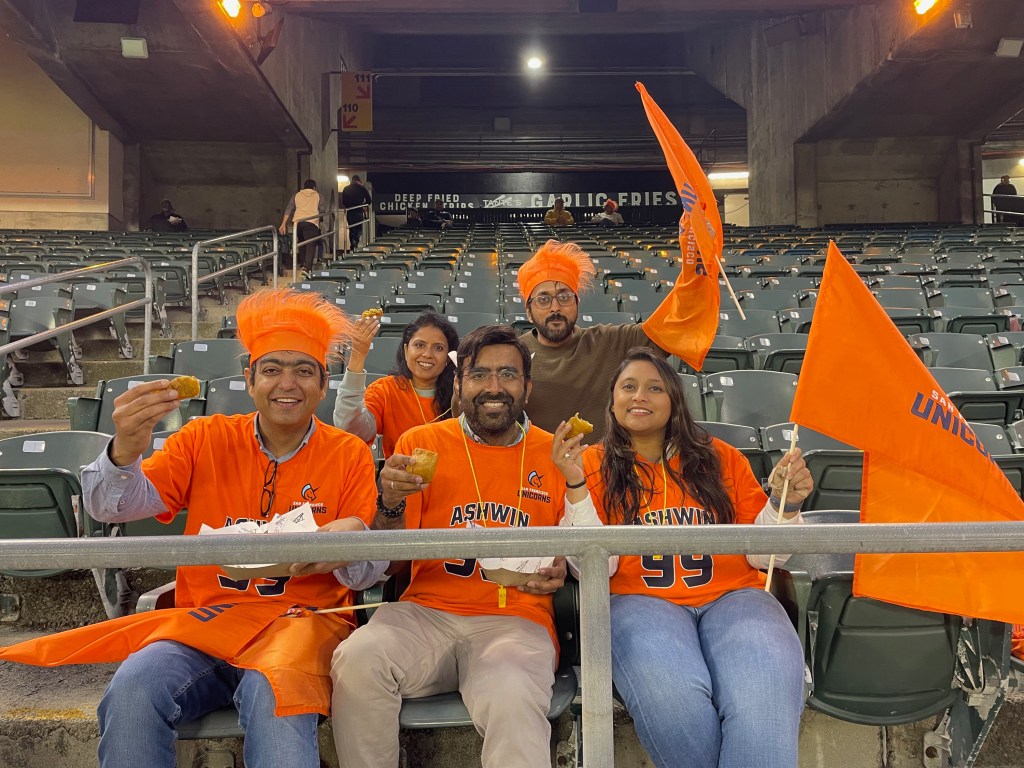 A group of four people wearing orange outfits and hats, holding flags and food, smile for a photo in an empty stadium seating area.