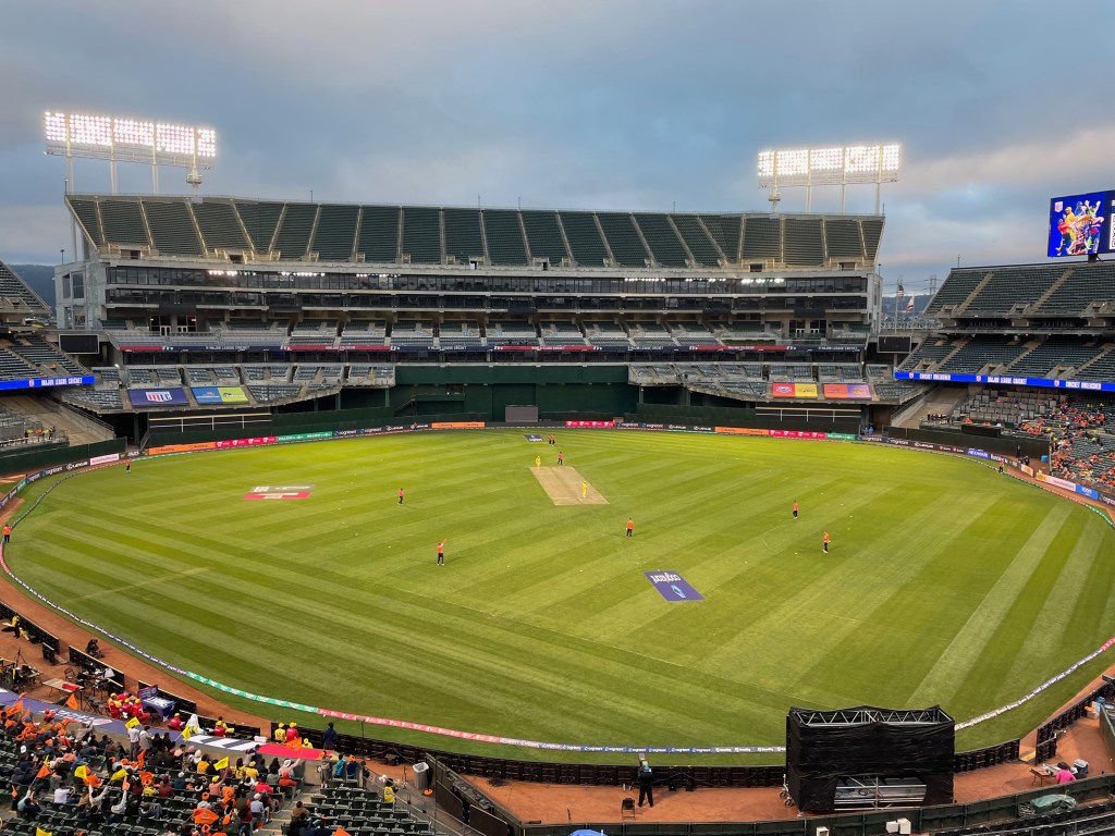 A cricket match is underway in a large, empty stadium with spectators in the stands and players on the field.