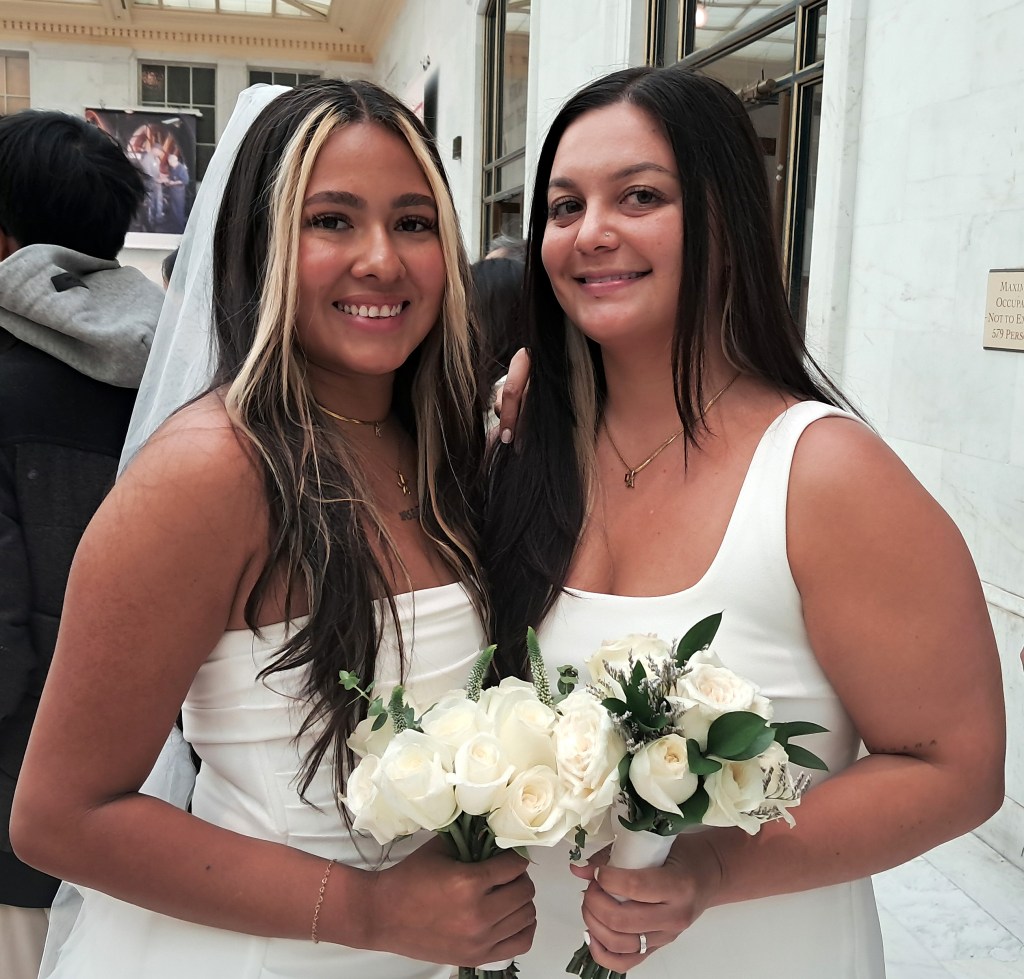 Two individuals in white dresses, one wearing a veil and holding a bouquet, smile together outdoors.