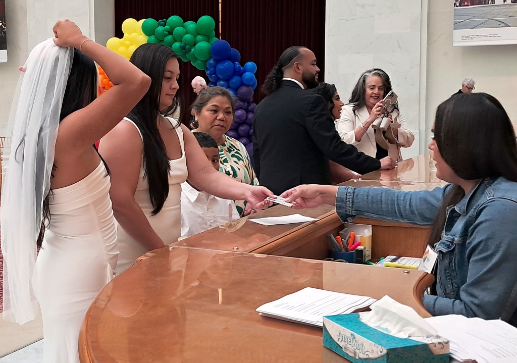A group of people, including a person in a white dress adjusting a veil, gather around a counter where another person is handing over an item to someone seated behind it, with colorful balloons in the background.