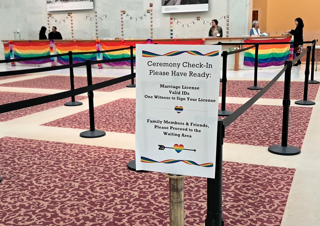 A sign at a wedding ceremony check-in area lists required documents and directs family members to the waiting area, with rainbow flags and “PRIDE” banners decorating the space.
