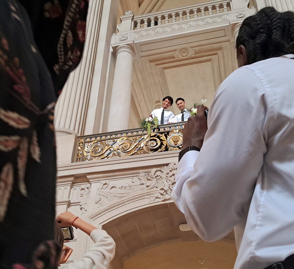 Two individuals stand on an ornate balcony in a grand hall, being photographed by people below.