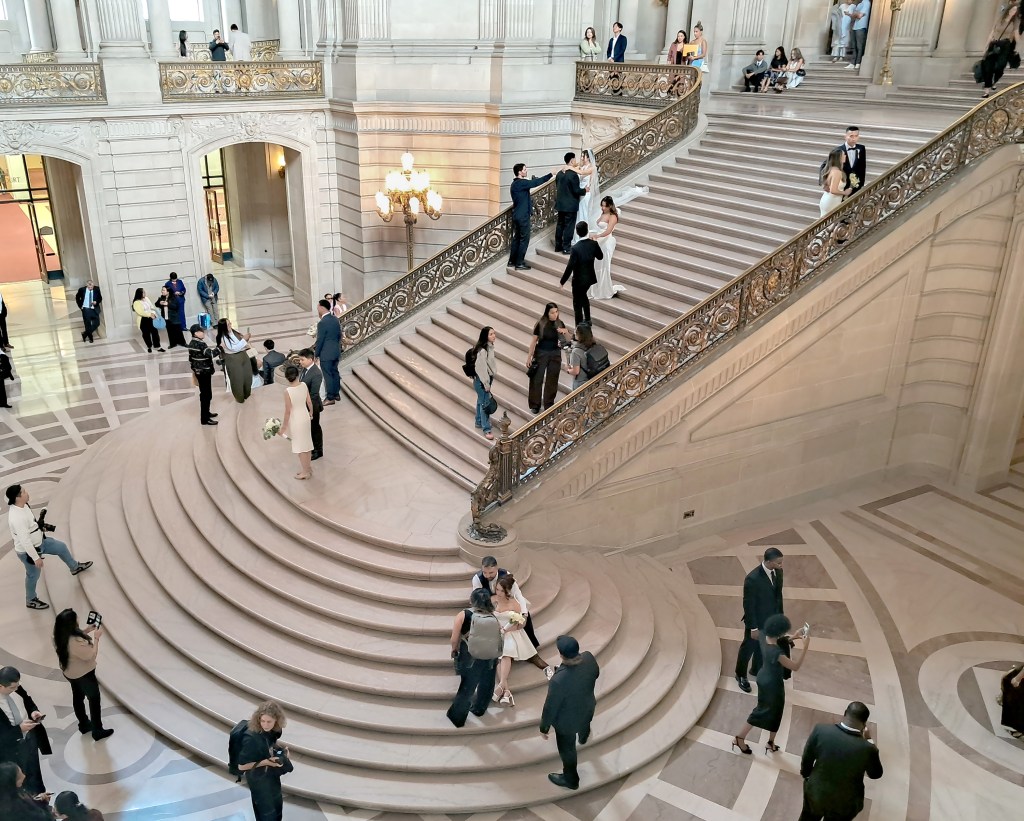 A couple walks down a grand, ornate staircase in a large, light-filled hall, surrounded by other people and photographers.