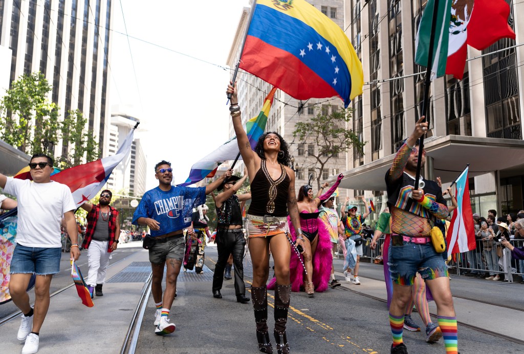 A vibrant parade features individuals proudly waving flags, including Venezuela’s, amidst urban skyscrapers and cheering crowds.
