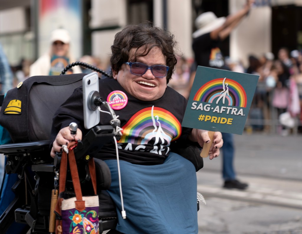 A person in a wheelchair smiles while holding a SAG-AFTRA Pride sign and wearing rainbow-themed apparel during a parade.