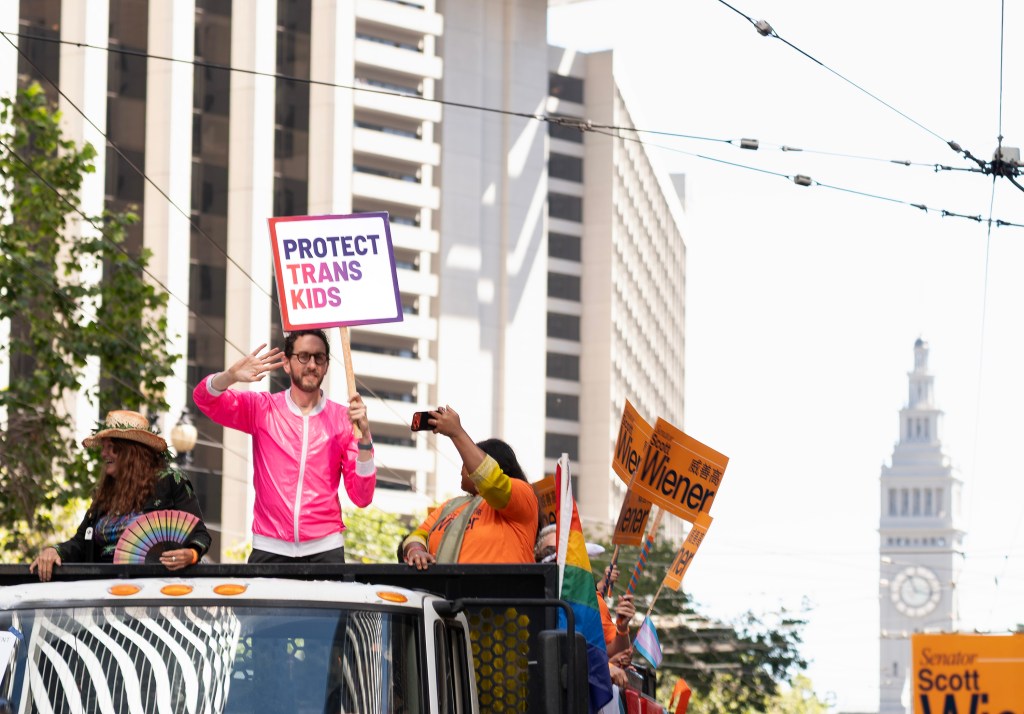 A person in a pink jacket waves from a parade float holding a sign that reads "PROTECT TRANS KIDS," surrounded by others holding signs and flags in an urban setting.