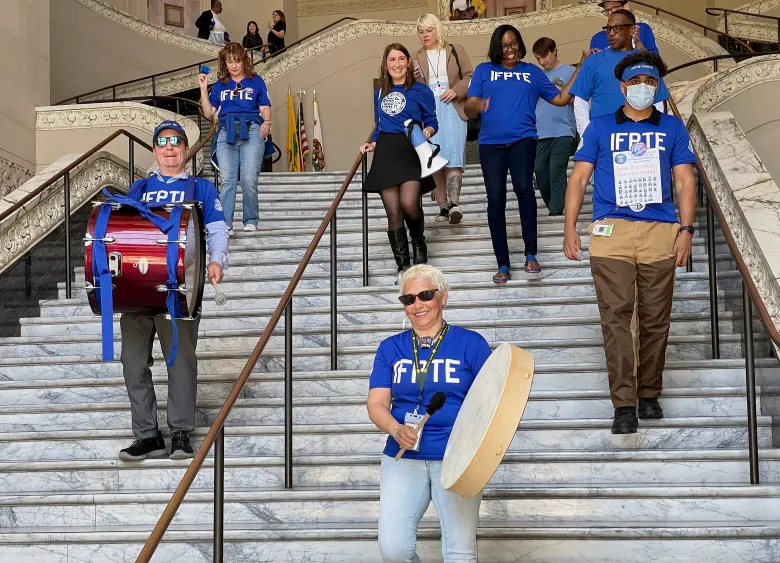 A group of individuals wearing blue IFPTE shirts march down a grand marble staircase, some carrying drums and percussion instruments.