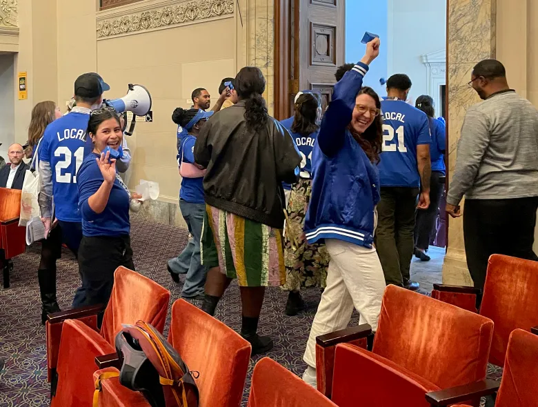 A group of people in blue jackets with "LOCAL 21" printed on them celebrate inside a formal building, some holding blue objects and a megaphone, surrounded by red seats.