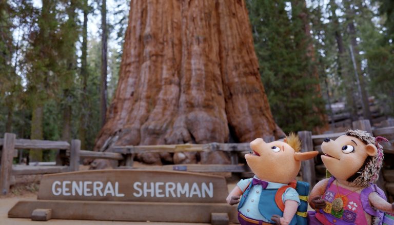 A screenshot from Wowsabout showing puppets Ronald and Roxy wearing backpacks and looking up in awe at the massive trunk of the General Sherman giant sequoia tree. A wooden sign reading