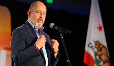 A man in a suit speaks into a microphone with a California state flag visible in the background.