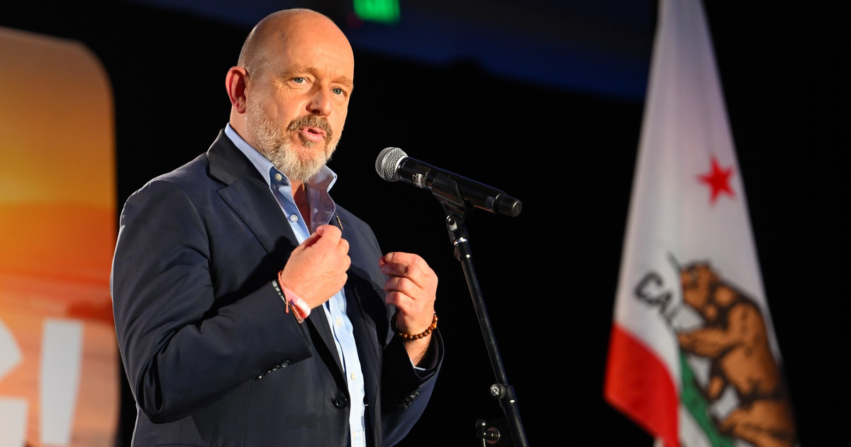 A man in a suit speaks into a microphone with a California state flag visible in the background.