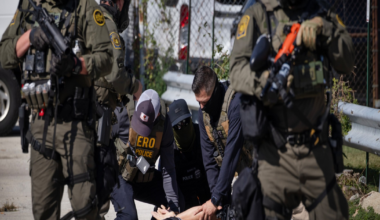 Several armed officers in tactical gear detain a person lying face down on the ground outside near a chain-link fence and parked vehicles.