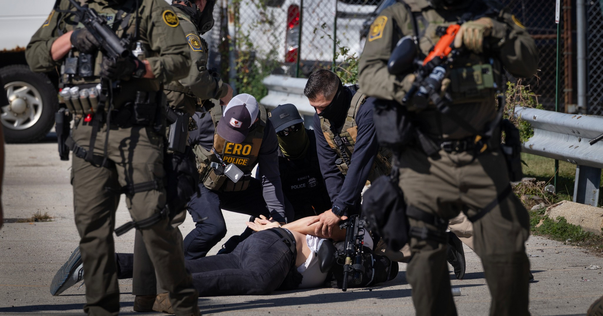 Several armed officers in tactical gear detain a person lying face down on the ground outside near a chain-link fence and parked vehicles.