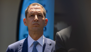 A man in a blue suit and patterned blue tie stands outdoors, looking serious, with part of another person and an NBC microphone visible nearby.