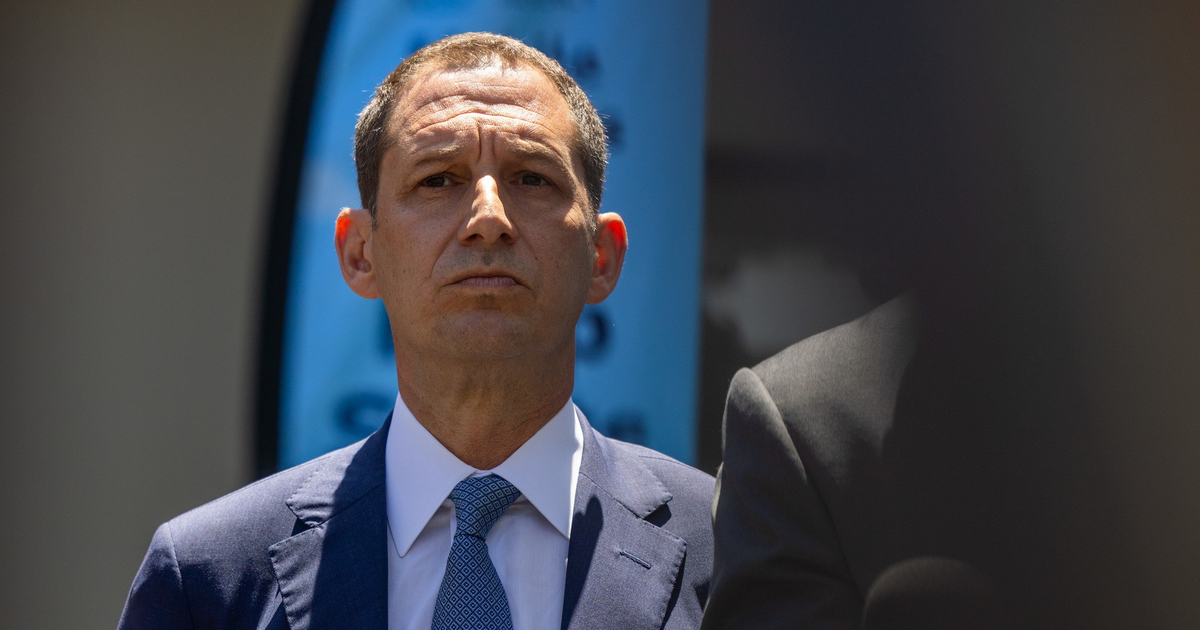 A man in a blue suit and patterned blue tie stands outdoors, looking serious, with part of another person and an NBC microphone visible nearby.
