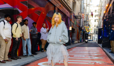 A young woman with blonde hair stands confidently in a layered skirt and oversized hoodie on a colorful urban street with onlookers around.