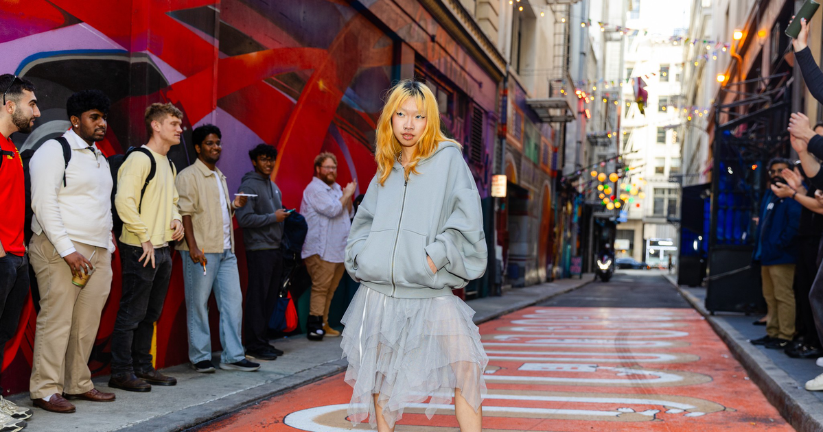 A young woman with blonde hair stands confidently in a layered skirt and oversized hoodie on a colorful urban street with onlookers around.