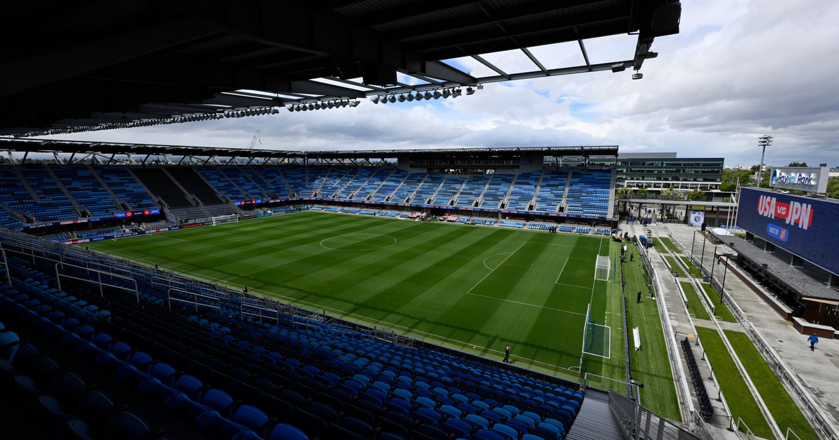 A large, empty soccer stadium with blue seats surrounds a green field, and a scoreboard nearby shows "USA vs JPN."