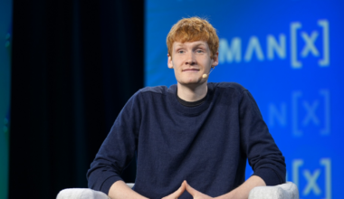 A young man with red hair, wearing a dark blue sweater, sits on a light gray chair with hands clasped and a microphone on his cheek.