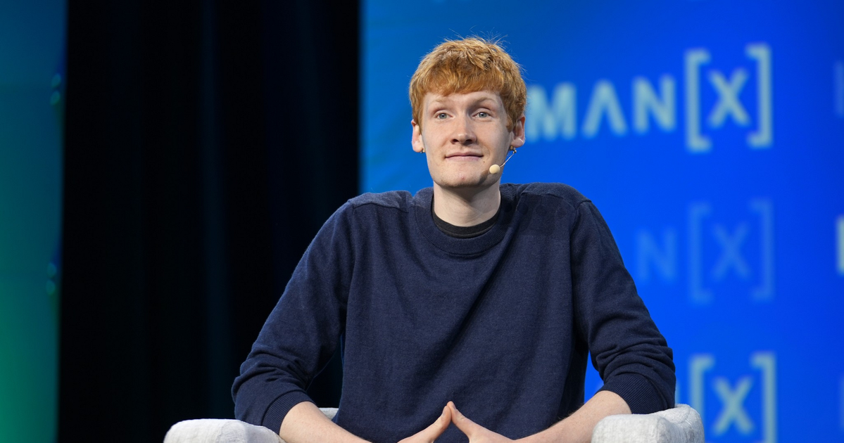 A young man with red hair, wearing a dark blue sweater, sits on a light gray chair with hands clasped and a microphone on his cheek.