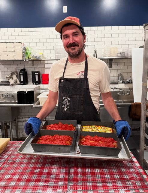 A man wearing an apron and blue gloves holds a tray with four loaf pans of food, standing in a commercial kitchen with tiled walls and stainless steel sinks.