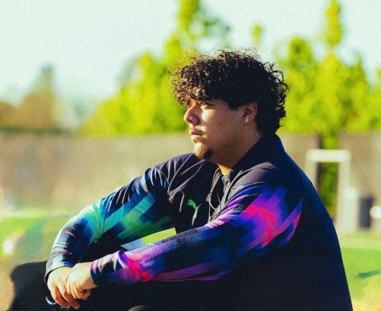 A person with curly hair and a beard sits outdoors, wearing a dark jacket with colorful geometric sleeves, looking to the left. Green trees are blurred in the background.