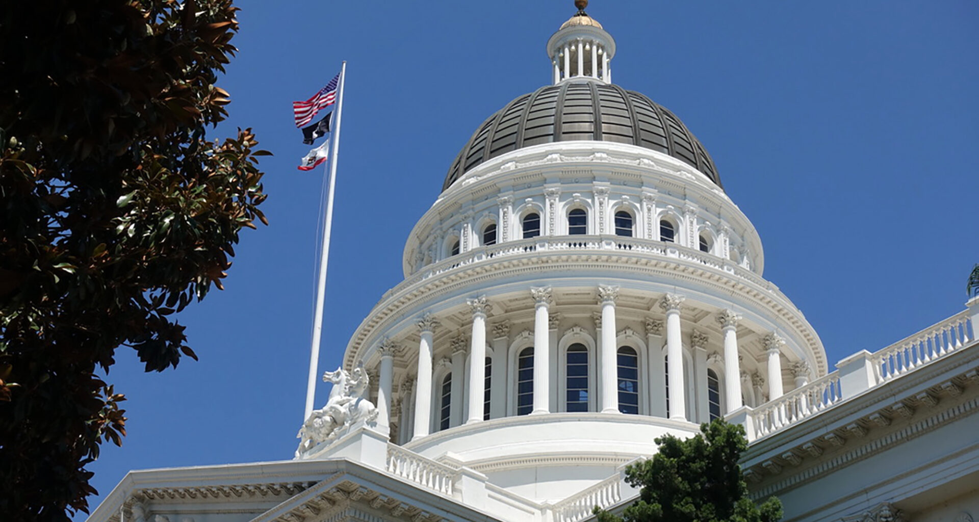 The dome of the California Capitol building in Sacramento, against a clear blue sky, with U.S. and state flags on the left.