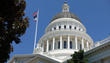 The dome of the California Capitol building in Sacramento, against a clear blue sky, with U.S. and state flags on the left.