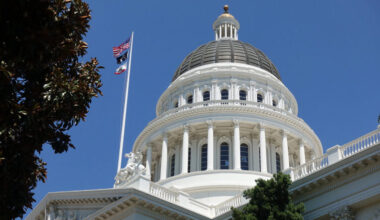 The dome of the California Capitol building in Sacramento, against a clear blue sky, with U.S. and state flags on the left.