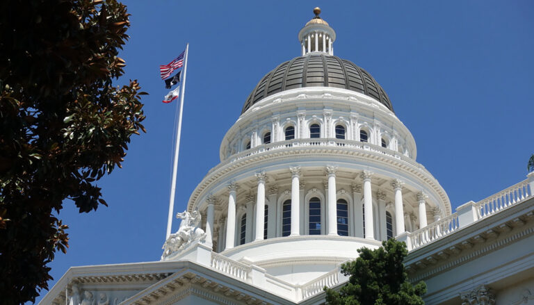 The dome of the California Capitol building in Sacramento, against a clear blue sky, with U.S. and state flags on the left.
