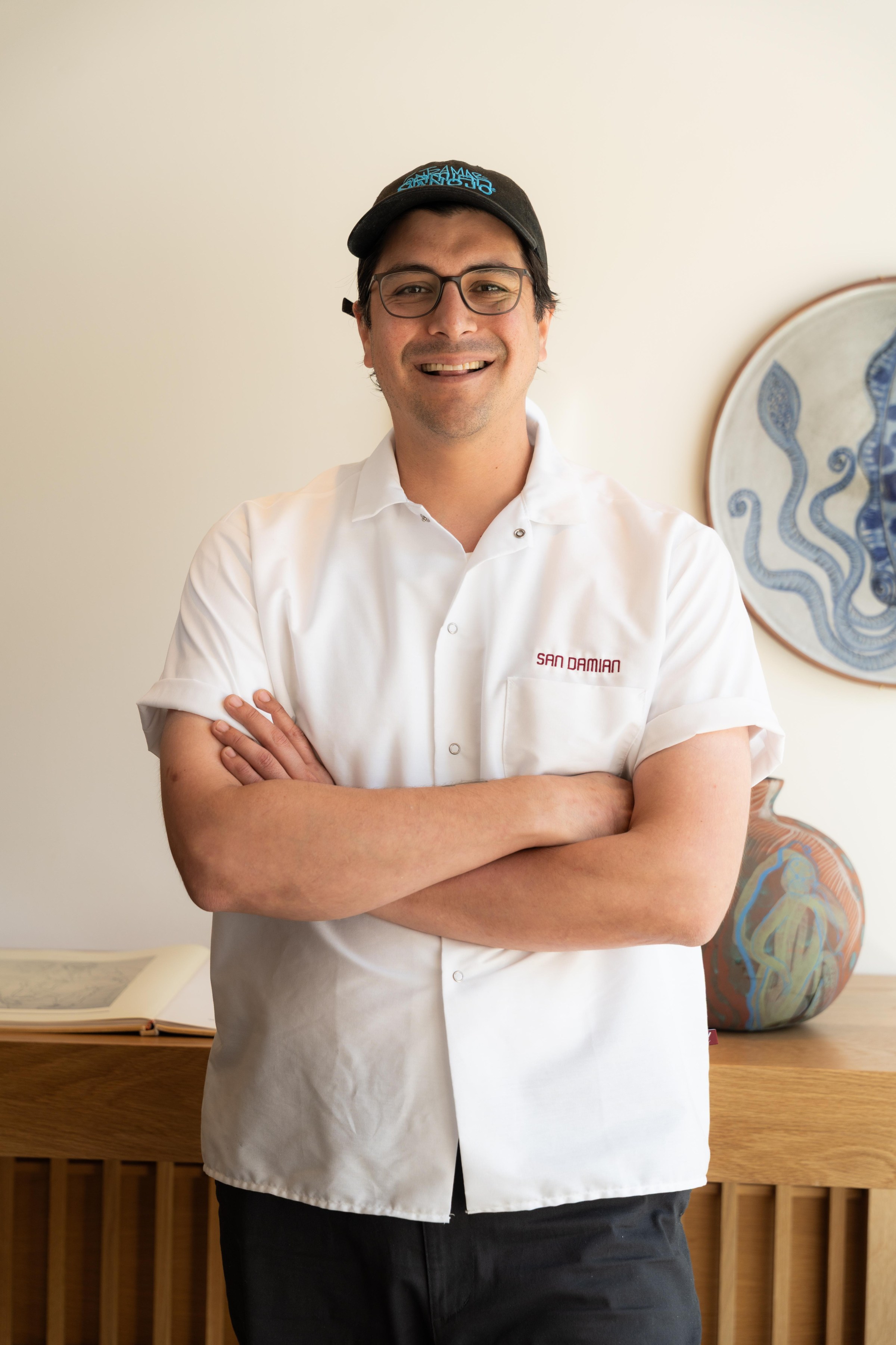 Chuy Cervantes, chef at San Damian, stands in a white chef’s jacket with San Damian embroidered on it.