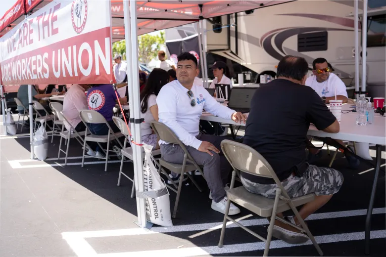 People seated under a tent at a union event, with a red and white "Iron Workers Union" banner, engaged in conversation and activities. Tone is collaborative.
