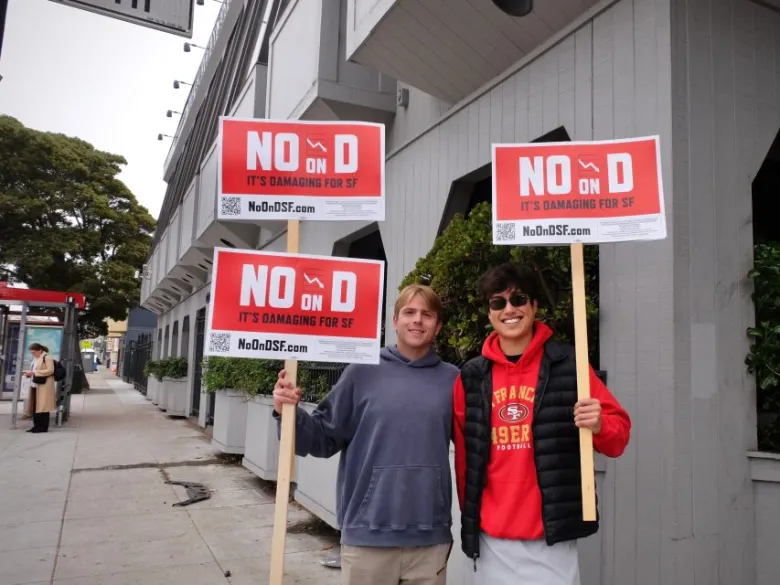 Two people stand on a sidewalk holding signs that read "No on D, It's damaging for SF" in front of a grey building, voicing their concerns ahead of the upcoming San Francisco election.