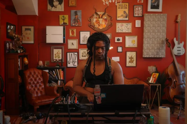A person with braided hair sits at a laptop in a red room decorated with framed art, musical instruments, and bookshelves.