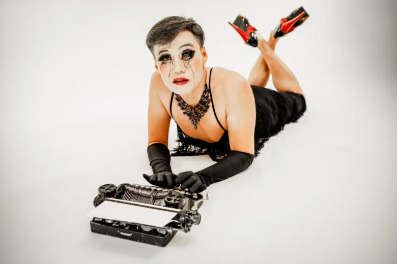 Person wearing dramatic makeup, black dress, gloves, and high heels lies on stomach in front of a vintage typewriter on a white background.