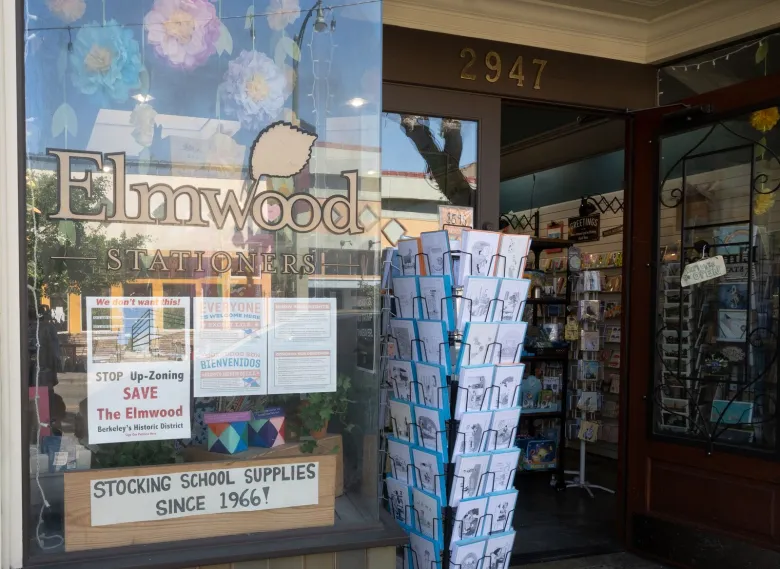A sign that reads "stop upzoning, save the Elmwood" is posted in the window of Elmwood Stationers, next to racks of cards
