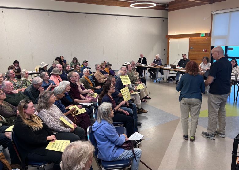 People fill a community meeting, many of them holding signs that read, "vote yes the Elmwood Historic District"