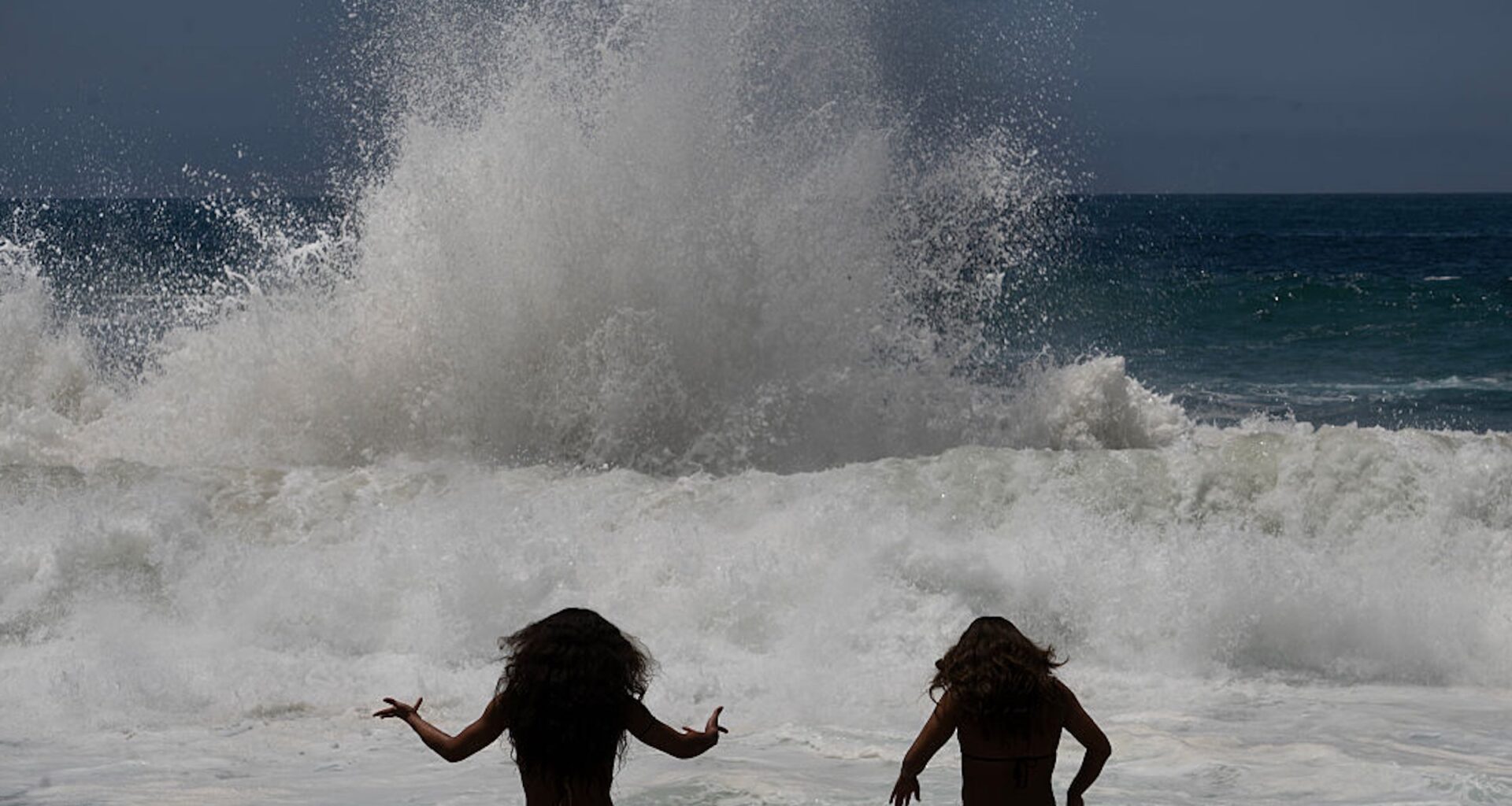 a silhouette of two young women is seen against the splashing of an ocean wave