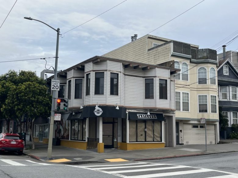 A two-story corner building with a sign reading "Tastetouse" stands quietly in Richmond. The windows are covered, and the street is empty except for a red car parked nearby.