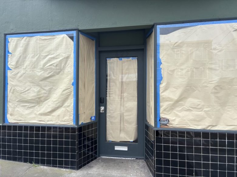 Storefront with windows and door covered in brown paper, blue painter's tape around edges, and black tile along the lower wall.