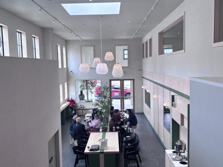 Modern cafe interior with high ceilings, pendant lights, and large windows. Groups of people are seated at tables, talking and eating. Bright natural light fills the space.