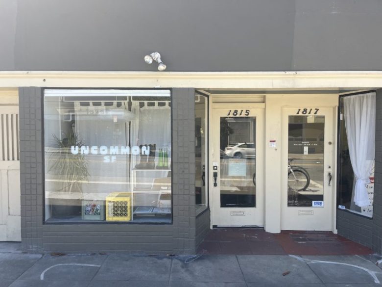 Street view of a storefront with a sign reading "UNCOMMON SF" in the window, located between doors numbered 1815 and 1817. A bicycle is visible through the glass door.