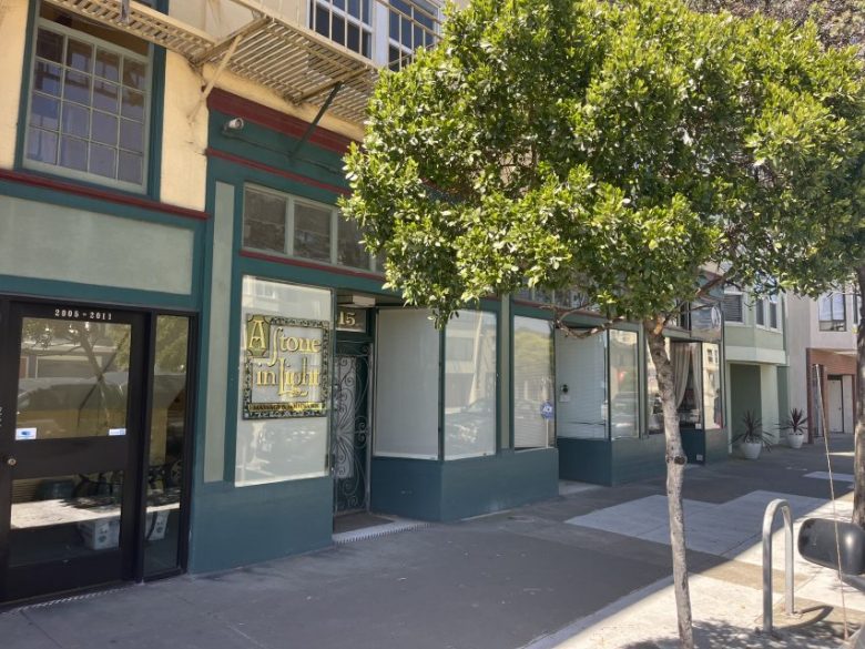 A sidewalk with a tree in front of storefronts, including one with a sign reading "A Stone in Light" near a door numbered 1515. Sunlight casts shadows on the scene.
