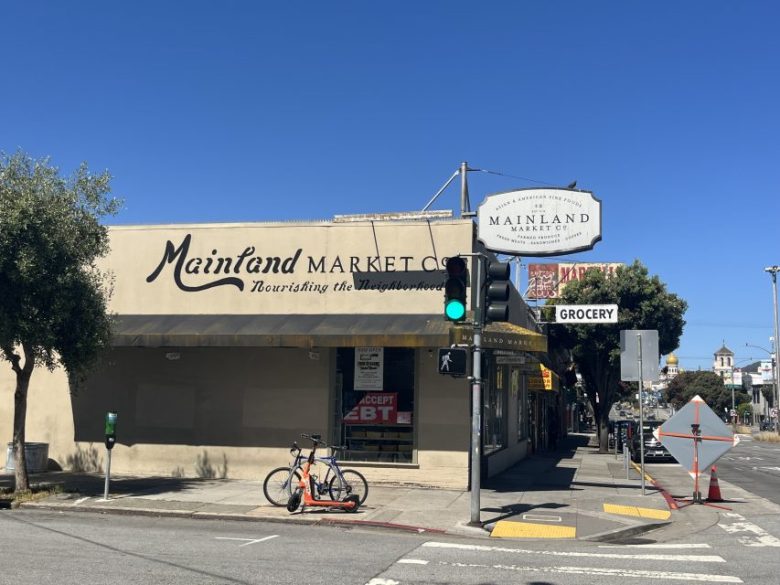A corner grocery store called Mainland Market Co. sits at an intersection with a bike parked outside and a green traffic light overhead on a sunny day.