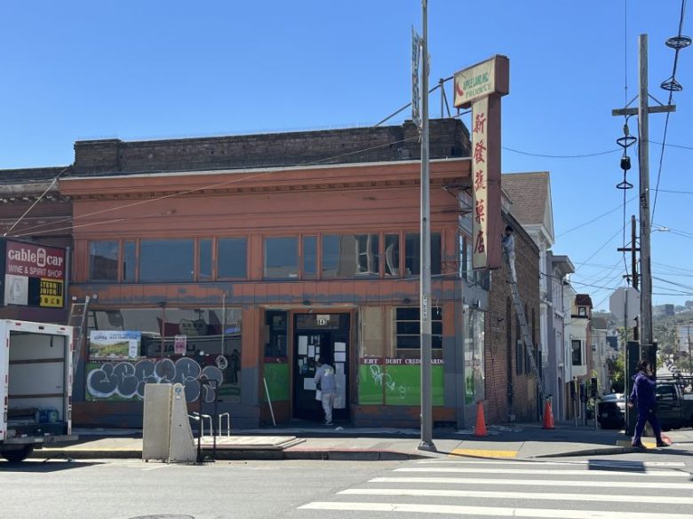 A brick corner store with red trim, a vertical sign with Chinese characters, and people entering. A truck is parked outside under a clear blue sky.