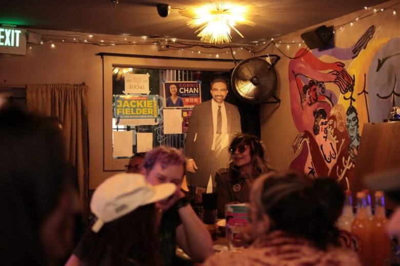 A group of people talk at a dimly lit bar with campaign posters mentioning tax reform and a cardboard cutout of a man on the wall behind them. A wall mural and string lights decorate the space.