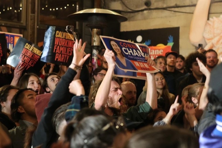 A crowd of people at an indoor event hold up political signs and cheer; some signs read “California Working Families Party” and “CONNIE CHAN FOR SUPERVISOR.”.