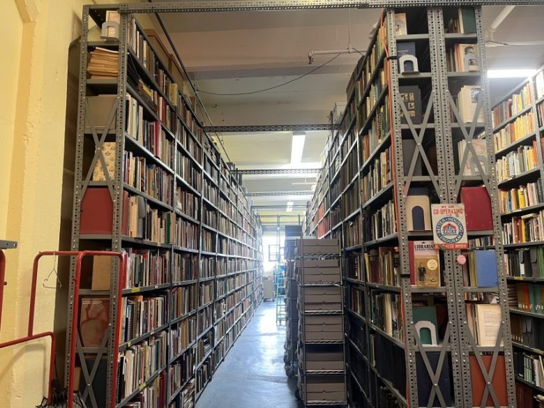 Aisle between tall metal bookshelves filled with books and storage boxes in a well-lit library or archive room.