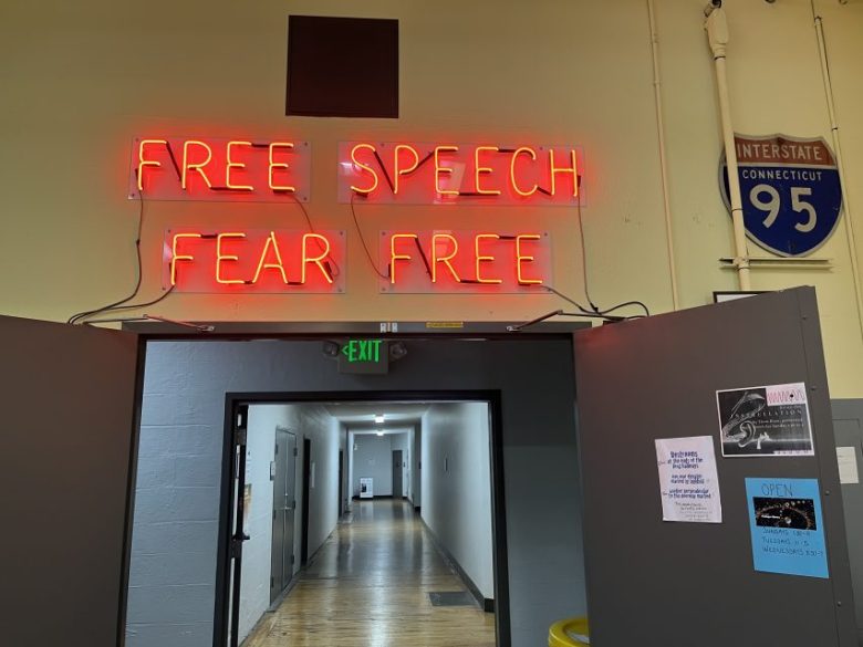 A neon sign reads "FREE SPEECH FEAR FREE" above double doors in a hallway, with an Interstate 95 Connecticut sign visible on the wall.