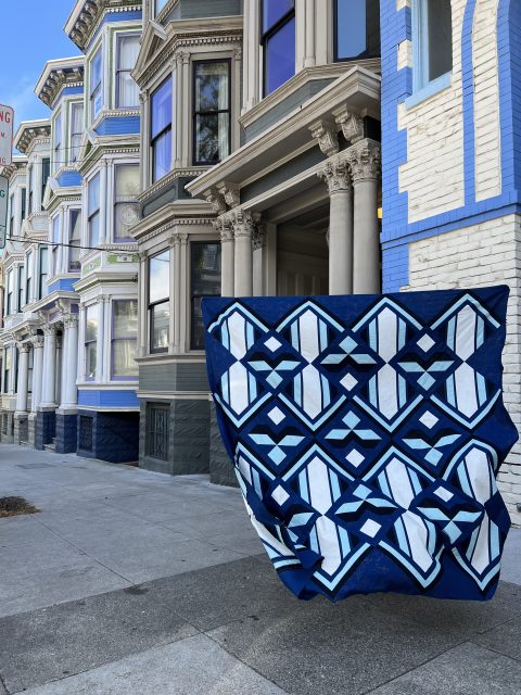 A blue and white geometric patterned quilt hangs in front of a row of ornate, Victorian-style houses on a city sidewalk.